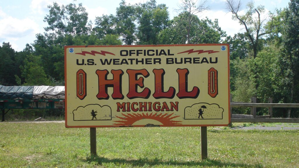 Official U.S. Weather Bureau sign in Hell, Michigan with bold red lettering, humorous graphics, and surrounded by trees on a sunny summer day.